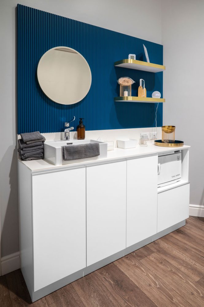 Un elegante bagno presenta un mobile bianco con lavabo moderno, asciugamani grigi, accessori in vetro e decorazioni minimaliste su una parete blu.