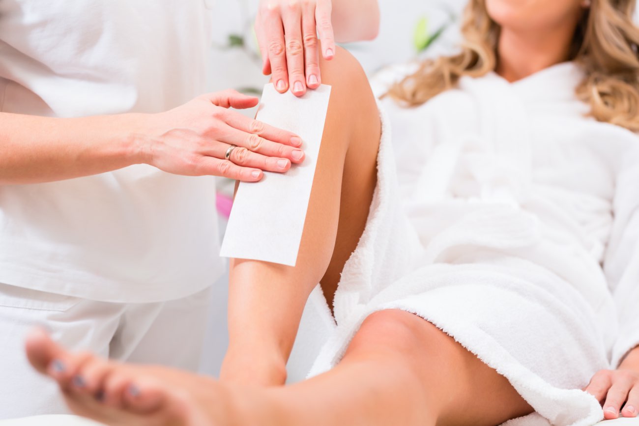 Una donna in accappatoio riceve un trattamento di epilazione artistica su una gamba da un'estetista. Ambientazione rilassante e professionale.