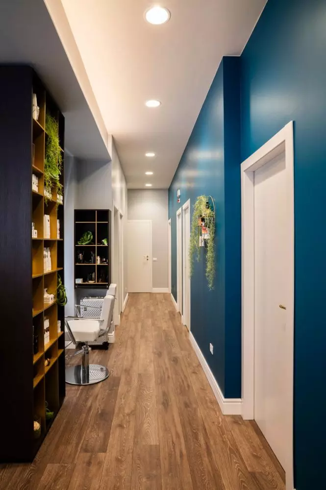 Un corridoio moderno di un centro estetico, con pareti blu e decorazioni verdi, scaffali contenenti prodotti di bellezza. Sul pavimento in legno chiaro, si scorge una poltrona bianca e porte chiuse ai lati.