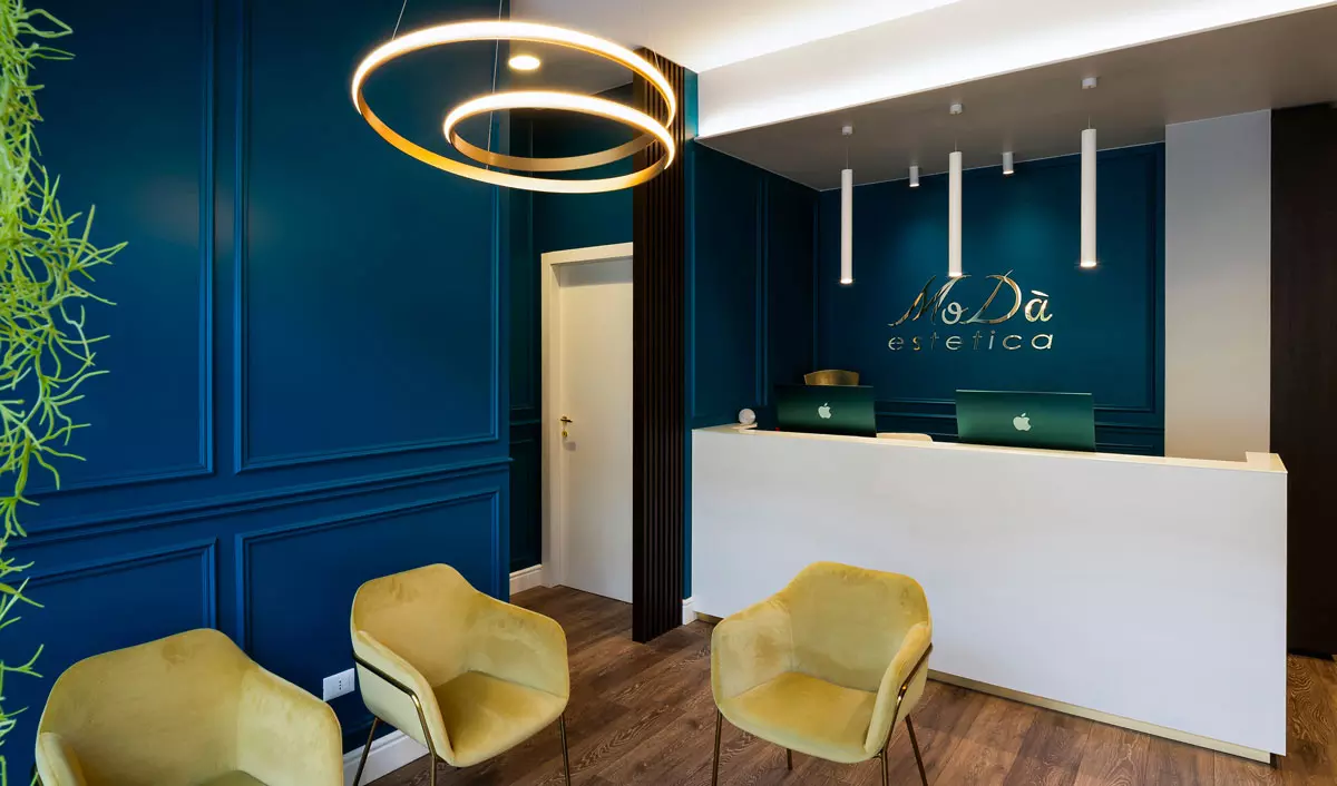 Interno di un centro estetico con pareti blu scuro, una reception elegante con logo e sedie gialle moderne, illuminazione soffusa.