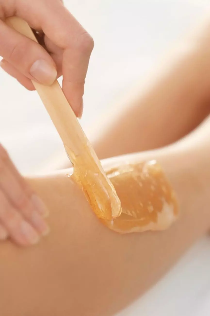 Un primo piano di una mano che applica una cera calda su un braccio, utilizzando una spatola in legno. La cera ha una consistenza dorata e lucida.