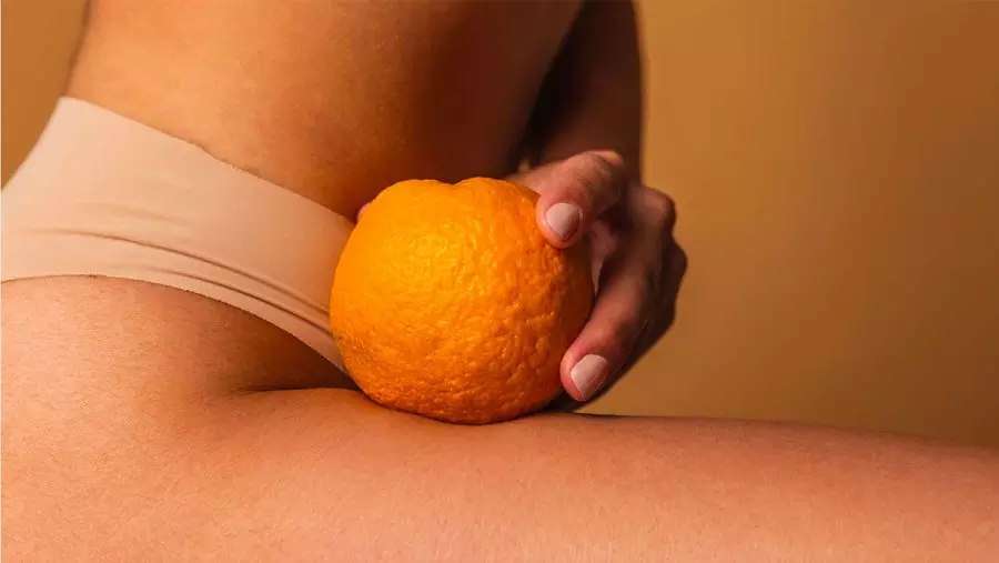 Una mano tiene un'arancia posta su una coscia nuda, in un contesto di colori neutri e caldi, evocando sensualità.