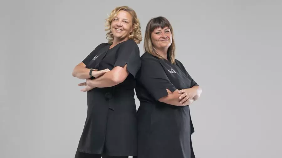 Monica e Daniela, sorridenti in abiti da lavoro neri, con le braccia incrociate, posano fianco a fianco su uno sfondo chiaro.