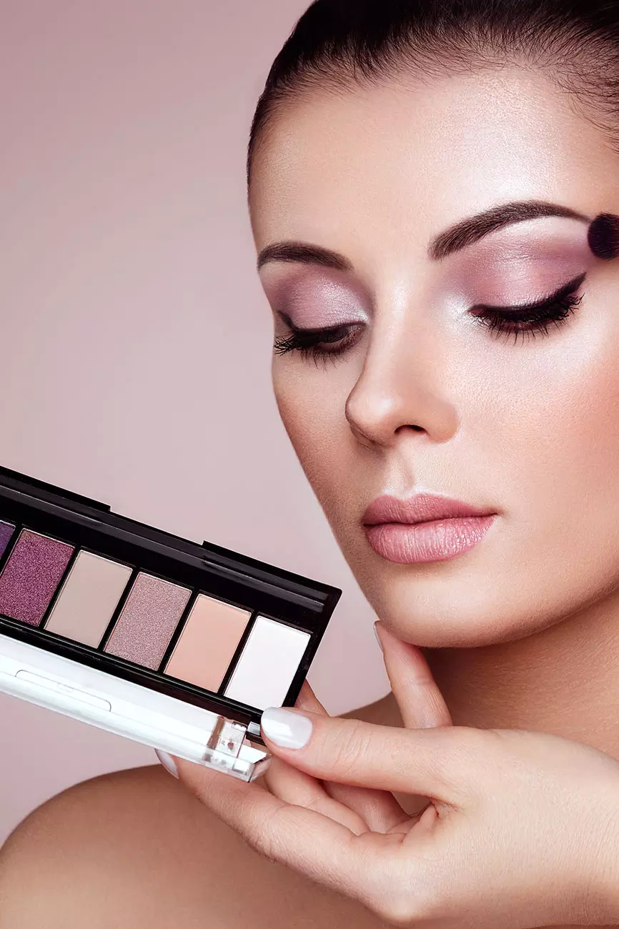 Primo piano di una donna con trucco elegante e palette di ombretti in mano, sfondo neutro. I colori della palette includono tonalità di viola e nude.